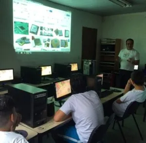 Grupo de pessoas sentadas em mesas, concentradas em uma tela à frente, em um ambiente de trabalho colaborativo.