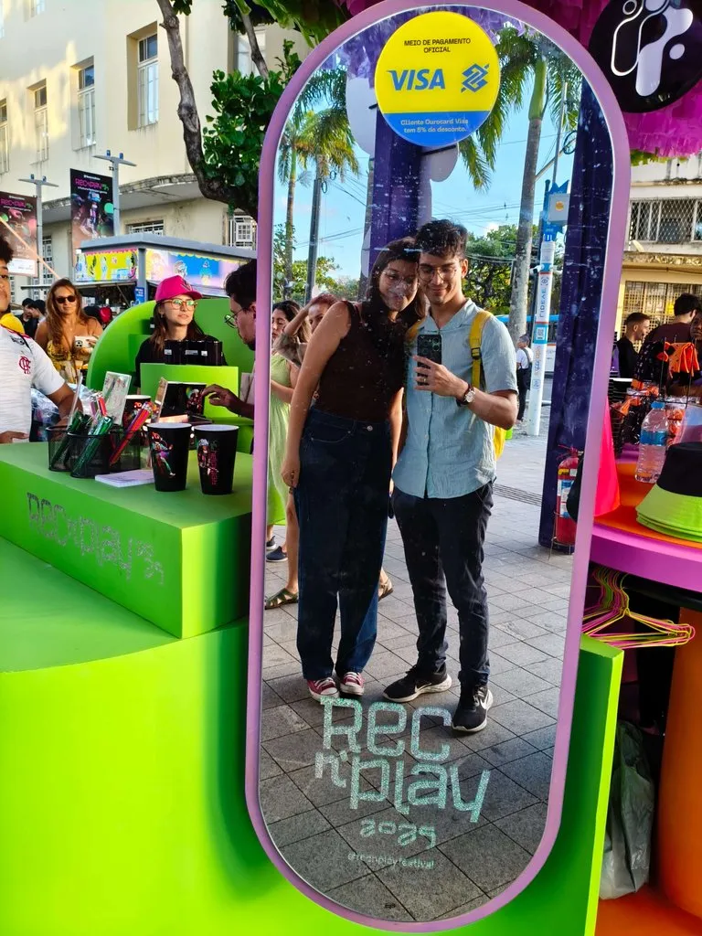 O autor do blog e sua namorada tirando uma selfie em um espelho com a marca 'Rec'n'Play 2025', em um evento de rua em Pernambuco, com barracas coloridas ao redor.