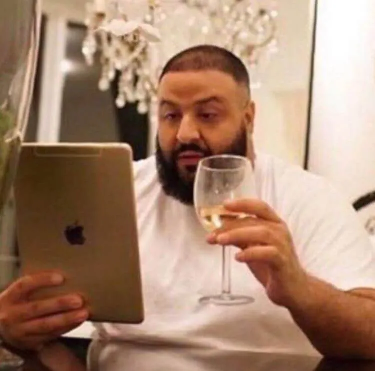 Meme do DJ Khaled sentado à mesa, segurando uma taça de vinho branco enquanto olha fixamente para um iPad