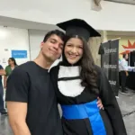 O autor do blog e sua namorada se abraçam e sorriem em uma foto durante a cerimônia de formatura. Ela veste a beca de graduação, capelo e faixa azul.