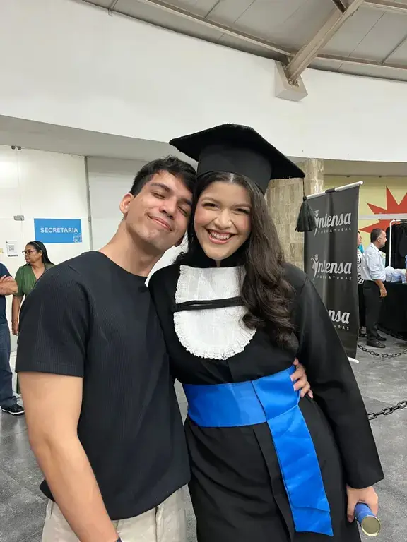 O autor do blog e sua namorada se abraçam e sorriem em uma foto durante a cerimônia de formatura. Ela veste a beca de graduação, capelo e faixa azul.