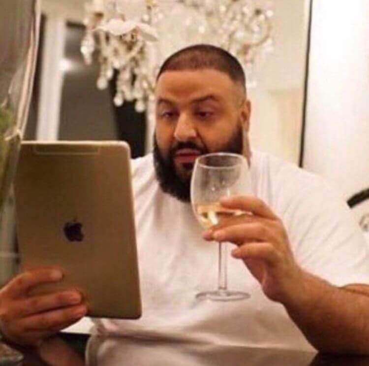 Meme do DJ Khaled sentado à mesa, segurando uma taça de vinho branco enquanto olha fixamente para um iPad