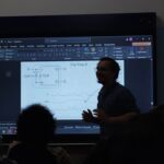 Foto da aula de organização de computador. Professor na frente do quadro, ao fundo, uma projeção mostra o diagrama de um Flip-Flop D e seu respectivo gráfico de temporização (clock).