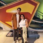 Fotografia do autor do blog e sua mãe sorridentes, posando em frente a um enorme totem iluminado com o icônico escudo "S" do Superman em um hall de cinema. Ele veste uma camisa aberta cor de abóbora sobre uma camiseta branca, e ela usa uma blusa branca e calça preta.