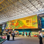 Fotografia de um grande pavilhão com teto de estrutura metálica. No centro, destaca-se um mural artístico em tons de amarelo e verde mostrando um mergulhador com escafandro antigo deitado e lendo um livro intitulado "Equilíbrio". Abaixo do mural, diversos visitantes caminham pelo pavilhão da Bienal.