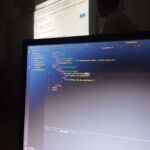 Close-up de um monitor exibindo o editor VS Code com um código HTML e JavaScript que verifica se os cookies estão habilitados. Ao fundo, a sala de aula da UERJ está obscurecida.