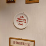 Fotografia de uma parede branca decorada. Ao centro, um prato de louça pendurado exibe a frase em vermelho: "todo abraço tem um pouco de Brasil".