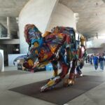 Fotografia de uma grande escultura colorida representando um lobo-guará, instalada no vão central da Cidade das Artes durante o Rio2C. A obra é inteiramente composta por pedaços de lixo plástico reciclado em tons de vermelho, azul, amarelo e laranja. Ao fundo, visitantes do evento caminham pela estrutura de concreto moderna do local.