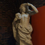 Fotografia de uma escultura clássica em mármore branco dentro de um museu. A estátua representa um jovem com cabelos cacheados, sem camisa e envolto em um manto, segurando uma máscara de teatro (tragedia) próxima ao ombro. O fundo é composto por uma parede de tijolos rústicos e um painel laranja vibrante à direita.