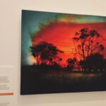 Fotografia de uma obra de arte em um museu. A imagem mostra silhuetas de árvores contra um céu vermelho intenso e dramático, com intervenções de tinta que sugerem fogo e fumaça. Ao lado, há um painel informativo com o título "Um Lugar Perdido" e o nome da artista.