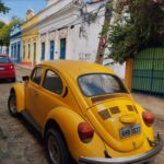 Fotografia de um Fusca amarelo clássico estacionado em uma rua de paralelepípedos. Ao fundo, veem-se casas históricas coloridas em tons de branco, amarelo e azul sob um céu limpo.