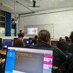 Estudantes em um laboratório de informática da UERJ. Em primeiro plano, um monitor exibe um código em linguagem C. Ao fundo, o professor aponta para uma projeção na lousa.