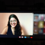 Fotografia de um monitor de computador exibindo uma videochamada. No centro da tela, Laura aparece sorridente, usando óculos e uma blusa preta, durante a defesa do seu Trabalho de Conclusão de Curso (TCC). À direita, veem-se as miniaturas desfocadas de outros participantes da reunião.