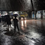 Fotografia vertical de uma rua urbana à noite sob chuva intensa. Em primeiro plano, a borda de um guarda-chuva preto enquadra a cena. No asfalto molhado e brilhante, refletem-se os faróis de um carro em movimento e as luzes dos estabelecimentos comerciais ao fundo. Pessoas com guarda-chuvas são vistas aguardando na calçada.