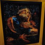 Fotografia de um quadro emoldurado em preto sobre uma parede amarela. A obra é uma colagem que forma o retrato de um homem de perfil, olhando para cima, com barba e cabelos em tons de dourado e cobre. As roupas são representadas por tons escuros e azuis, e o fundo é composto por pequenos recortes pretos com figuras humanas em miniatura.