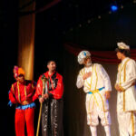 Fotografia de uma cena teatral no palco do Teatro dos Grandes Atores. O fundo é escuro com cortinas vermelhas discretas.