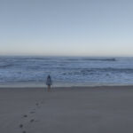 Fotografia em tons suaves de cinza e azul de uma praia deserta. Uma pessoa caminha de costas em direção ao mar, deixando uma trilha de pegadas nítidas na areia úmida. O horizonte é limpo e as ondas quebram suavemente na orla.