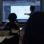 Vista da sala de aula na UERJ onde o professor aponta para uma tabela verdade de um Flip-Flop RS projetada em uma lousa digital. Alunos são vistos de costas em suas carteiras.