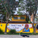 Fotografia em plano aberto da fachada da Casa Arte Vida. O muro amarelo agora exibe um grafite de uma criança sorridente com pinturas tribais no rosto ao lado da frase "Educar é semear o futuro!". À direita, o artista está em cima de uma cadeira pequena fazendo os últimos detalhes no muro. Árvores altas emolduram a cena.